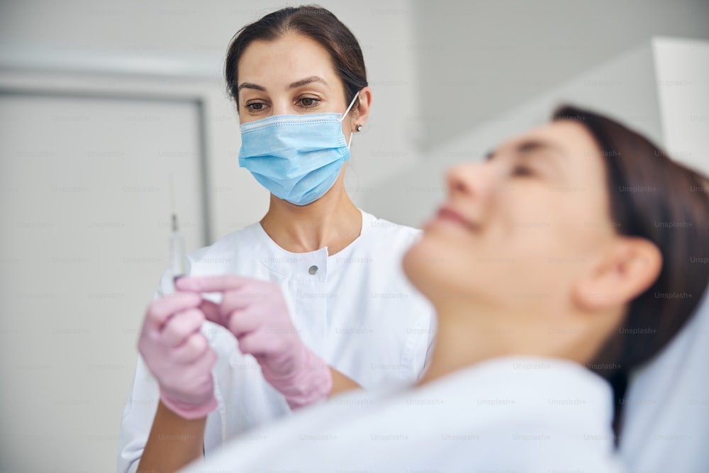 Concentrated female cosmetologist in a face mask preparing for injecting a smiling serene middle-aged woman