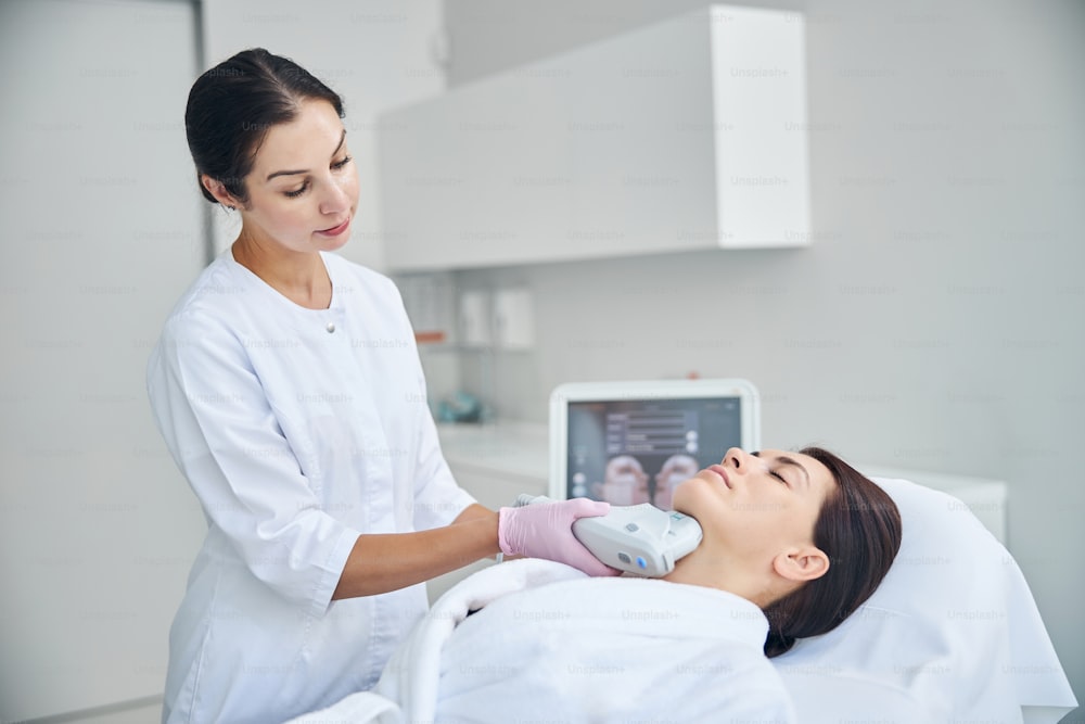Experienced female dermatologist using a beauty device for tightening the skin on her patient neck