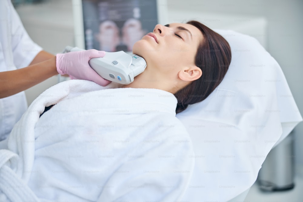Serene female patient with closed eyes getting an ultrasonic facial treatment performed by a cosmetologist