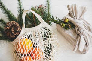 素朴な木製の背景に緑のスプルースとオレンジと緑の枝と再利用可能なショッピングバッグとリネン生地で包まれたスタイリッシュなクリスマスギフト。ゼロウェイストのクリスマス休暇