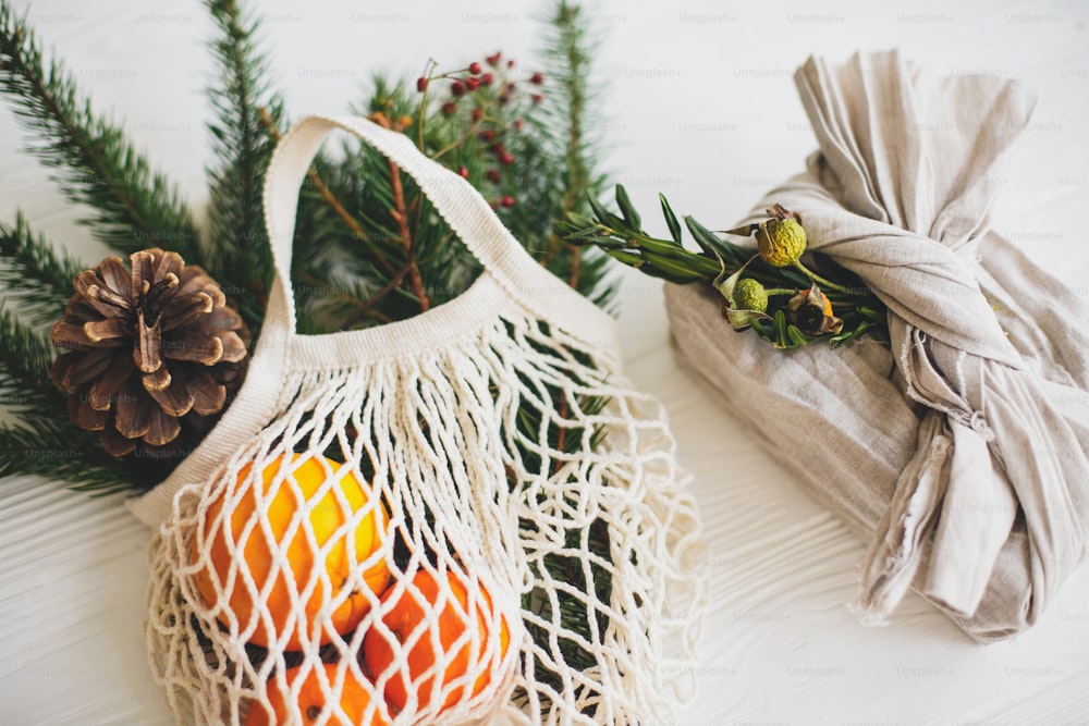 Stylish christmas gift wrapped in linen fabric with green branch and reusable shopping bag with green spruce and oranges on rustic wooden background. Zero waste christmas holidays