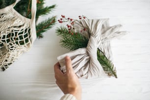 素朴な白い木製の背景に緑のモミの枝とリネン生地に包まれたスタイリッシュなクリスマスプレゼントを手に。ゼロウェイストのクリスマス休暇。サステナブルなライフスタイル