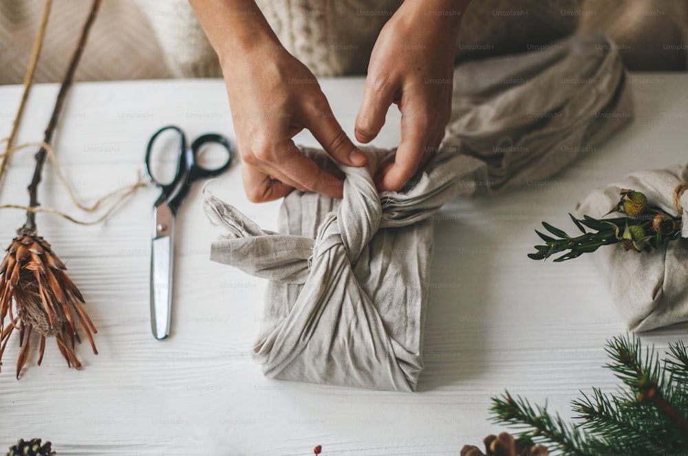 Hands wrapping christmas gift in linen fabric in furoshiki style on white rustic table with natural fir, herb, pine cones and scissors. Female preparing plastic free christmas present