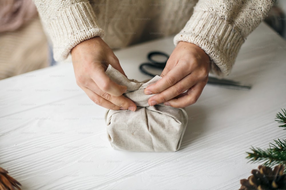 Woman in cozy sweater preparing plastic free christmas present, zero waste holidays. Female hands wrapping christmas gift in linen fabric on wooden table with green branch, pine cones, scissors.