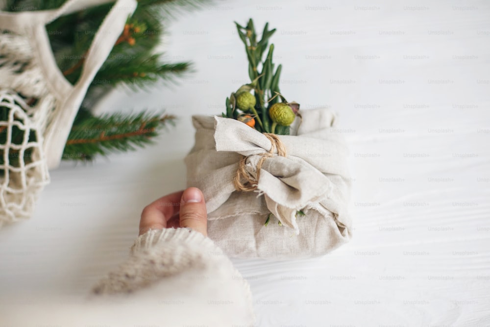 Hand holding stylish christmas gift wrapped in linen fabric with green  branch on rustic white wooden background. Zero waste christmas holidays. Sustainable lifestyle