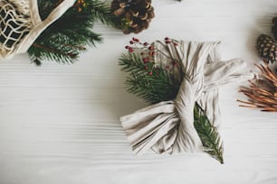 Stylish christmas gift flat lay. Present wrapped in linen fabric decorated with natural fir branch on white rustic table background with pine cones and herbs. Zero waste winter holidays
