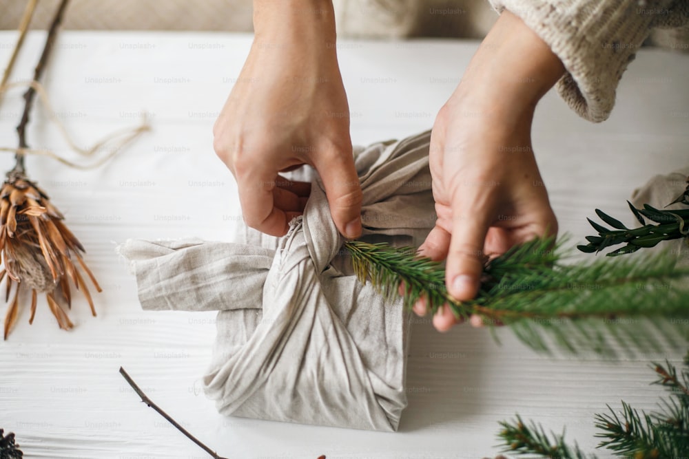 Mains décorant un cadeau de Noël élégant en tissu de lin avec une branche de sapin vert sur une table rustique blanche avec des pommes de pin et des baies. Fleuriste préparant un cadeau de Noël zéro déchet. Des vacances sans plastique