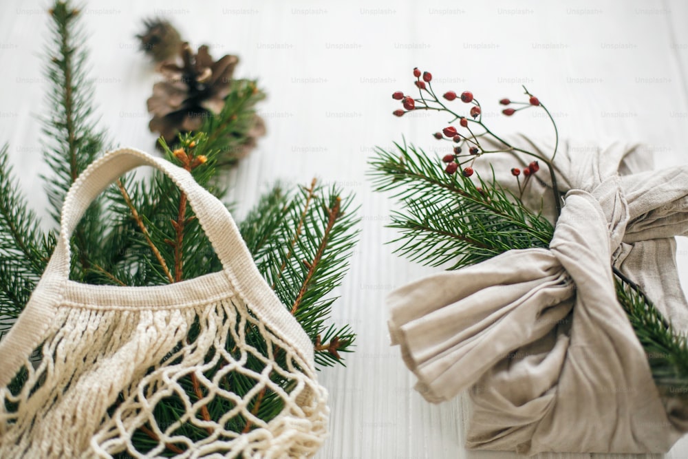 Stilvolles Weihnachtsgeschenk eingewickelt in Leinenstoff mit grünem Zweig und wiederverwendbarer Einkaufstasche mit grüner Fichte auf rustikalem Holzhintergrund. Zero Waste Weihnachtsferien. Nachhaltiger Lebensstil