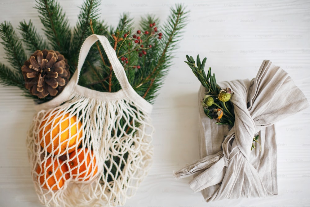 Zero waste christmas holidays. Stylish christmas gift wrapped in linen fabric with green branch and reusable shopping bag with green spruce and oranges on rustic wooden background