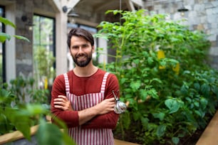 Portrait of man gardener with apron standing in greenhouse, looking at camera.
