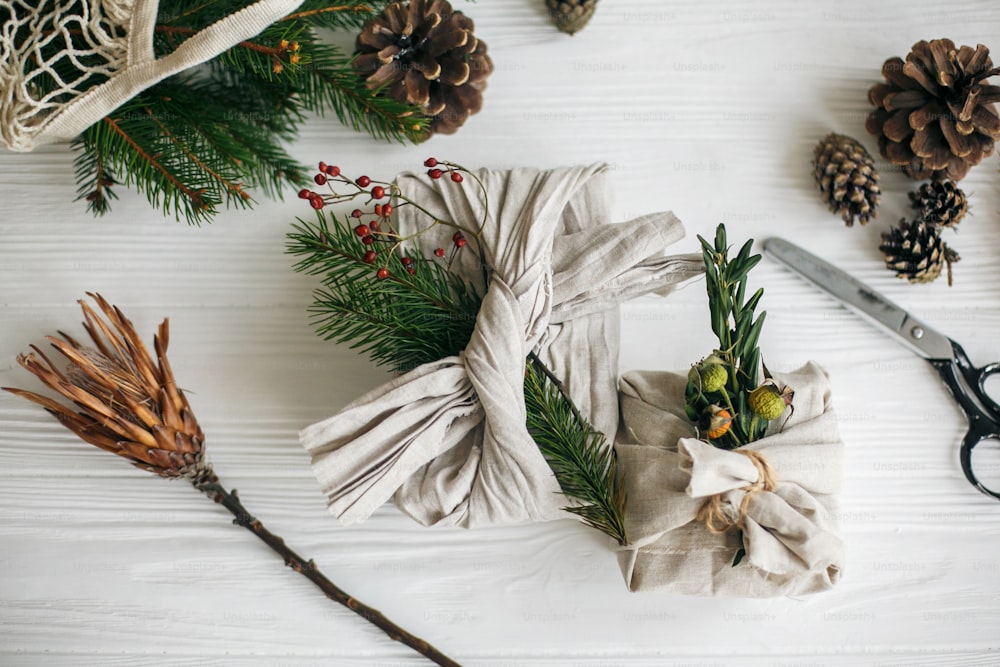 제로 웨이스트 세련 된 크리스마스 선물. 전나무와 가위가 있는 흰색 소박한 테이블 배경에 천연 녹색 가지가 있는 린넨 패브릭으로 포장된 선물. 플라스틱 무료 휴일.
