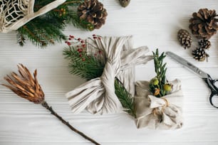 Zero waste stylish christmas gift. Gift wrapped in linen fabric with natural green branch on white rustic table background with fir and scissors. Plastic free holidays.