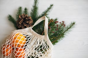 Net cotton bag with green spruce branches, oranges and pine cones on white rustic background. Reusable shopping bag with winter decorations, plastic free holidays. Top view