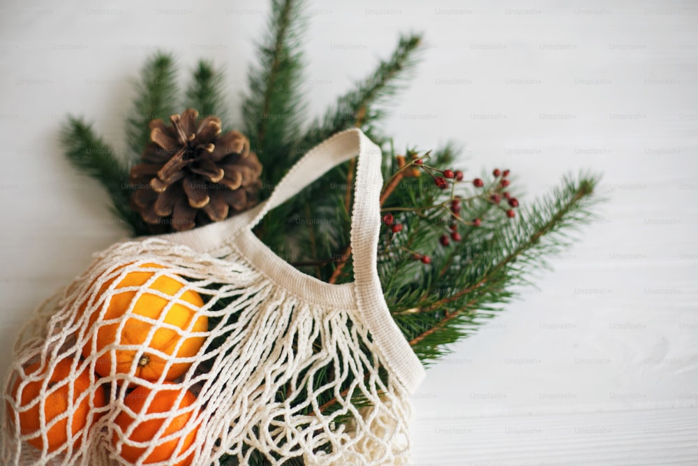 Net cotton bag with green spruce branches, oranges and pine cones on white rustic background. Reusable shopping bag with winter decorations, plastic free holidays. Top view