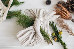 Stylish christmas gift flat lay. Present wrapped in linen fabric decorated with natural fir branch on white rustic table background with pine cones and herbs. Zero waste winter holidays