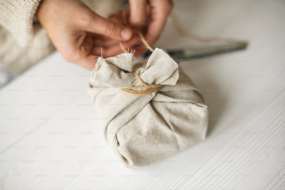 Hands wrapping stylish christmas gift in linen fabric on white rustic table with scissors. Female in cozy sweater preparing plastic free christmas present, zero waste holidays
