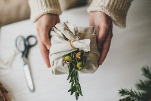 Zero Waste Weihnachtsgeschenk. Hände mit stilvollem Geschenk, eingewickelt in Leinenstoff mit naturgrünem Zweig auf rustikalem Hintergrund mit Tannenzapfen und Schere. Plastikfreier Urlaub