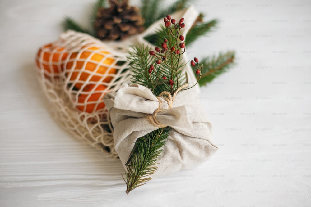 Stylish christmas gift wrapped in linen fabric with natural green branch and red berries on background of reusable cotton bag with oranges on white  table. Zero waste christmas holidays
