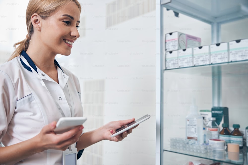 Jolly young woman in medical office standing near cabinet with medicine while using tablet and smartphone