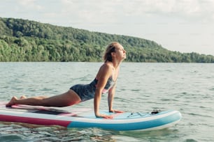 Middle age Caucasian woman practising yoga on paddle sup surfboard at sunset. Female stretching doing workout on lake water. Modern individual outdoor summer aquatic sport activity.