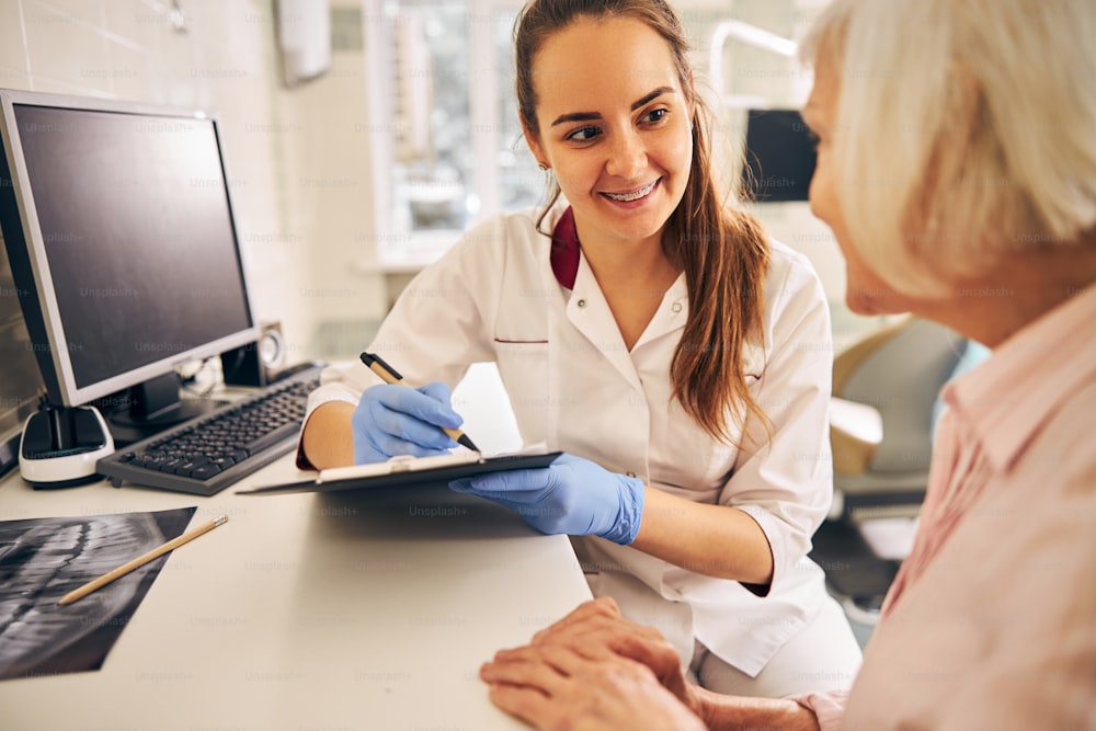 Waist up portrait of cute brunette doctor holding clipboard while listening patient and doing notes