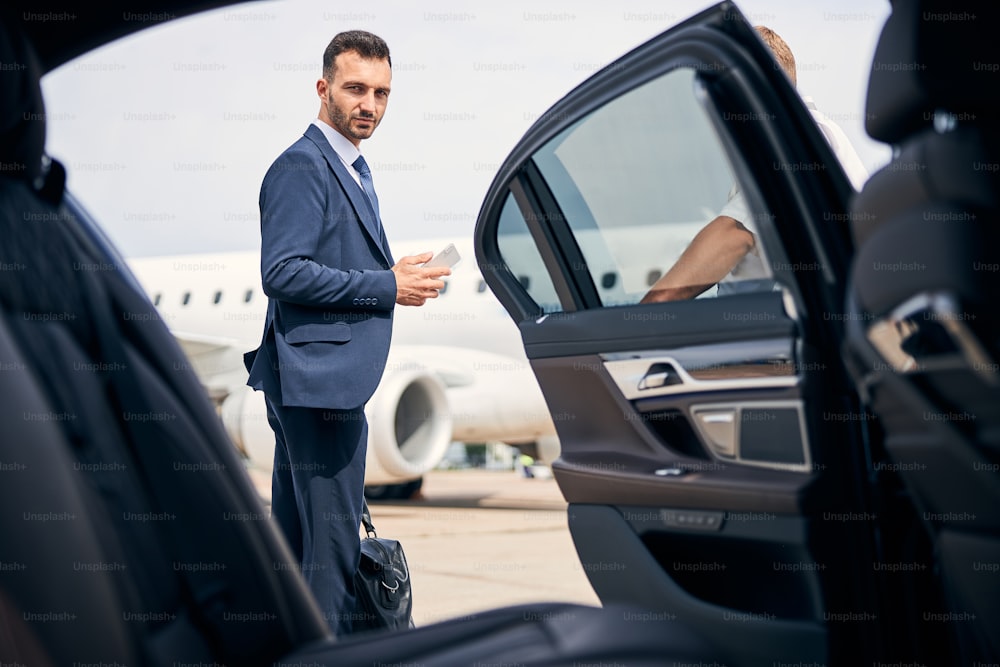 Handsome brunette man in a classy suit holding his carry-on standing by a taxi cab