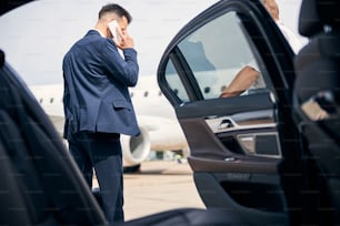 Joven hombre de negocios hablando por teléfono parado junto al taxi con una puerta abierta que lo recogen del vuelo