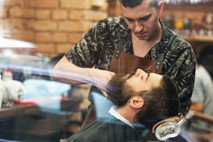 Bearded male sitting in an armchair in a barber shop while hairdresser trip his beard with scissors. Close-up of barber shearing beard to man in barbershop