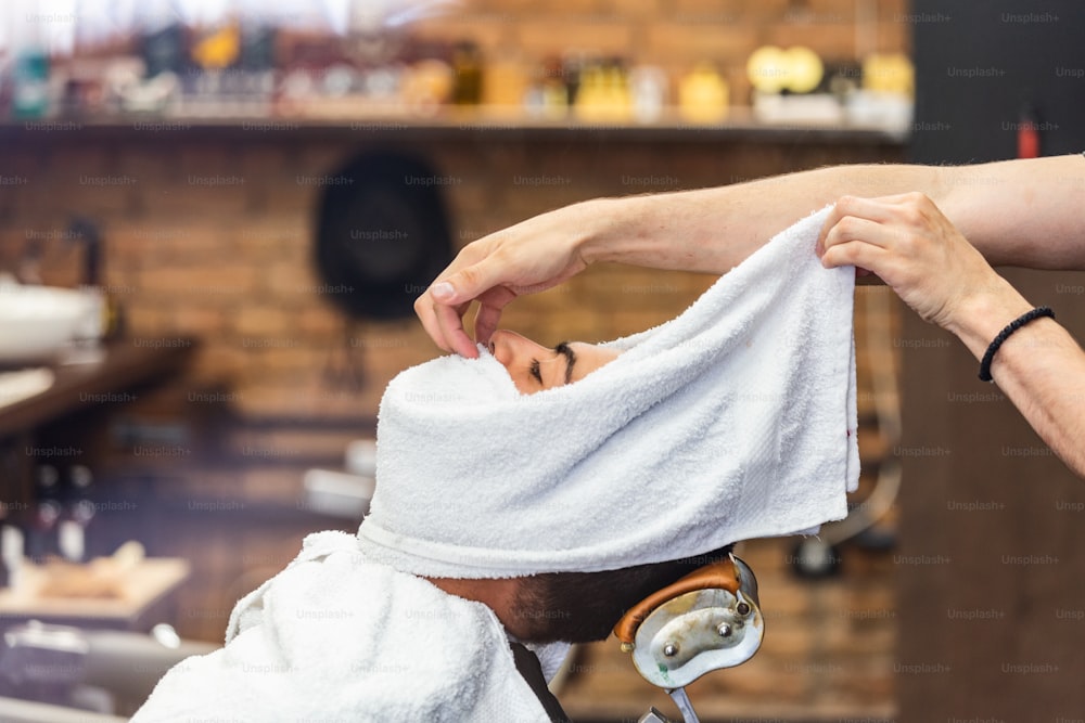 Barbier couvre le visage d’un homme avec une serviette chaude. Rituel traditionnel de rasage de la barbe avec des compresses chaudes et froides dans un salon de coiffure à l’ancienne. Client avec une serviette chaude sur le visage avant de se raser dans un salon de coiffure
