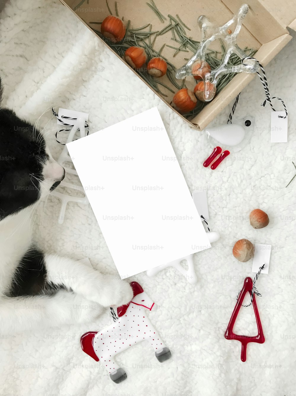 Christmas greeting card mockup, empty white postcard with space for text and cute cat paws holding modern festive glass ornaments on white soft blanket. Top view