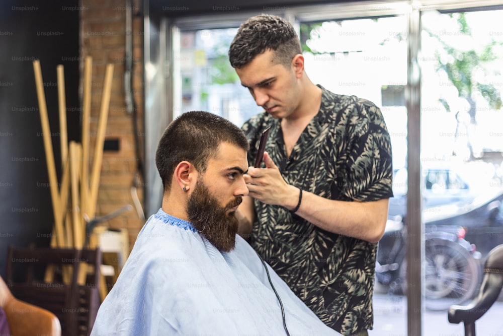 Rendre la coupe de cheveux parfaite. Jeune homme barbu se faisant couper les cheveux par un coiffeur alors qu’il est assis sur une chaise chez le coiffeur