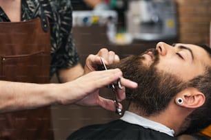 Hombre barbudo sentado en un sillón en una peluquería mientras el peluquero se tropieza la barba con unas tijeras. Primer plano del barbero cortando la barba al hombre en la barbería
