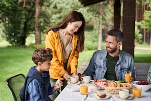 Charming young woman holding cup of tea and smiling while her husband and son sitting at the table with delicious food and drinks