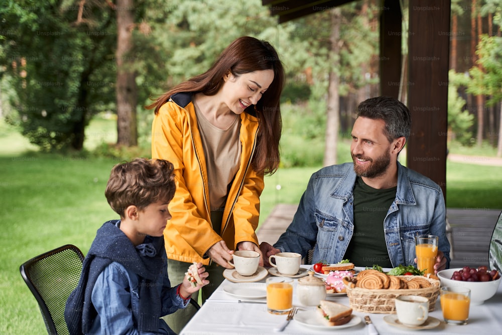 Charmante jeune femme tenant une tasse de thé et souriant tandis que son mari et son fils sont assis à la table avec de délicieux plats et boissons