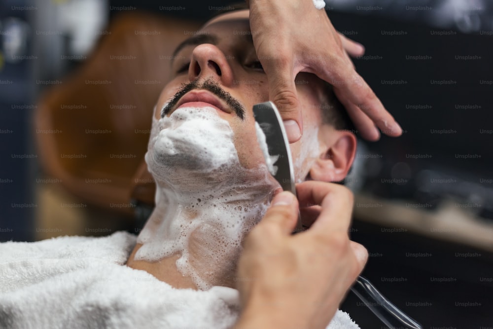 男の口ひげは剃っている。専門の理髪師の手にあるかみそり。理髪店で昔ながらの髭剃りをする青年。クローズアップ、レトロなスタイルの画像