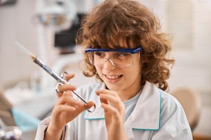 Close-up photo of a curly kid with a sneaky smile wearing a lab coat holding a medical syringe and looking cunning