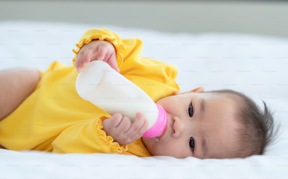 Bambino carino con il biberon sul letto, piccolo bambino asiatico tailandese sdraiato sul letto bianco che beve il latte dal biberon