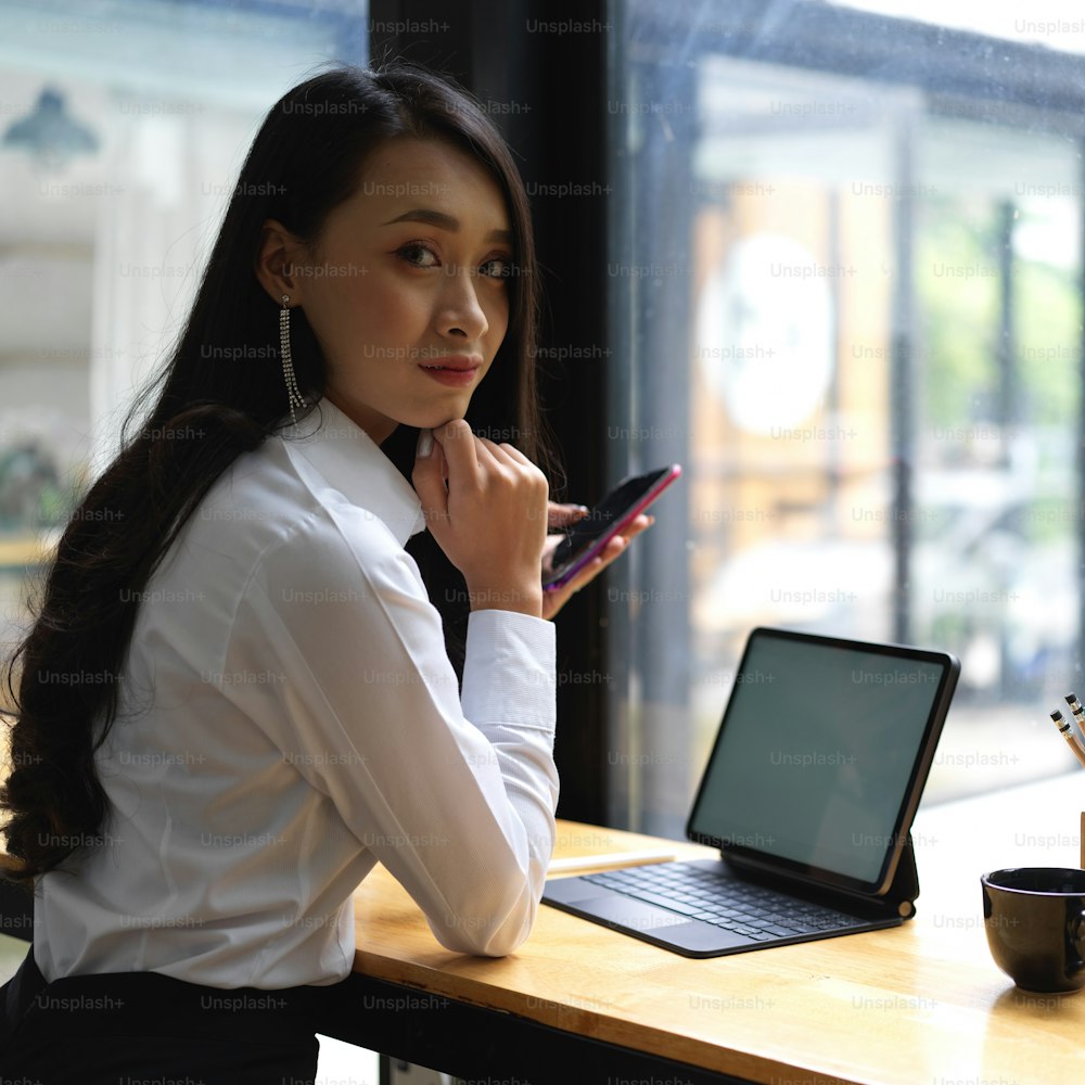 Portrait of female worker looking into camera while working with smartphone and tablet in cafe