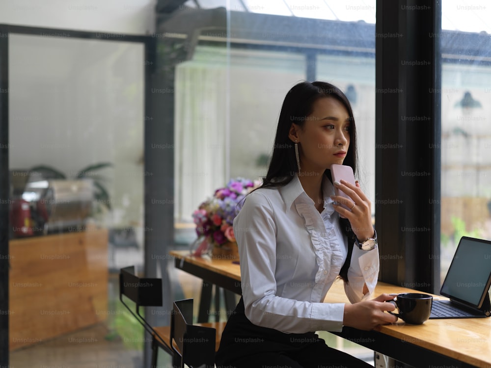 Portrait of thoughtful female holding smartphone and coffee cup while sitting at workplace