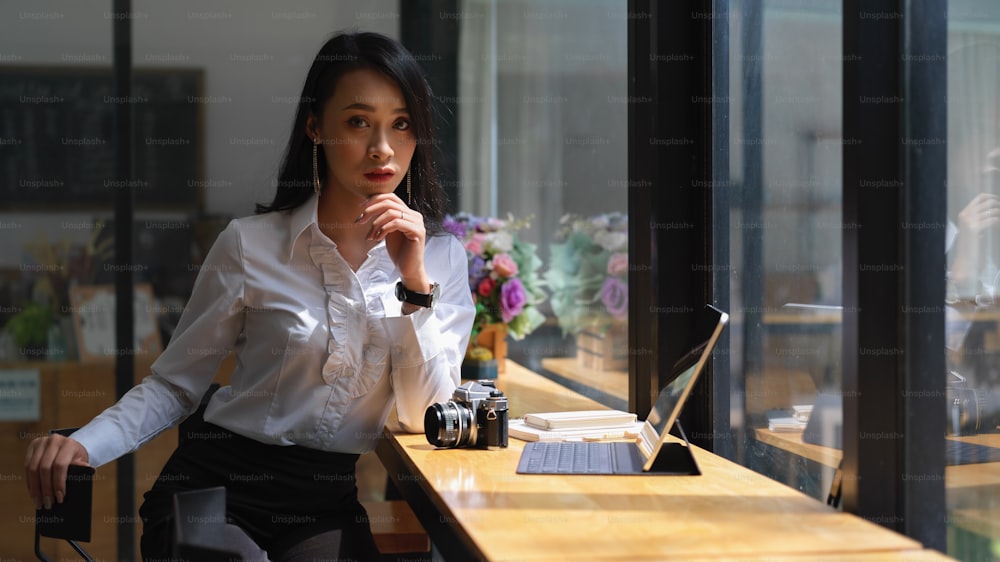 Ritratto di giovane e attraente freelance femminile seduto al bar di legno con fotocamera e tavoletta digitale nel caffè