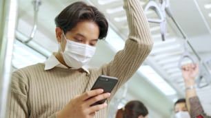 Traveler wearing face mask while using mobile phone on public train . Coronavirus disease or COVID 19 pandemic outbreak and urban city lifestyle problem in rush hour commuting concept .