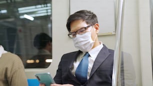 Traveler wearing face mask while using mobile phone on public train . Coronavirus disease or COVID 19 pandemic outbreak and urban city lifestyle problem in rush hour commuting concept .