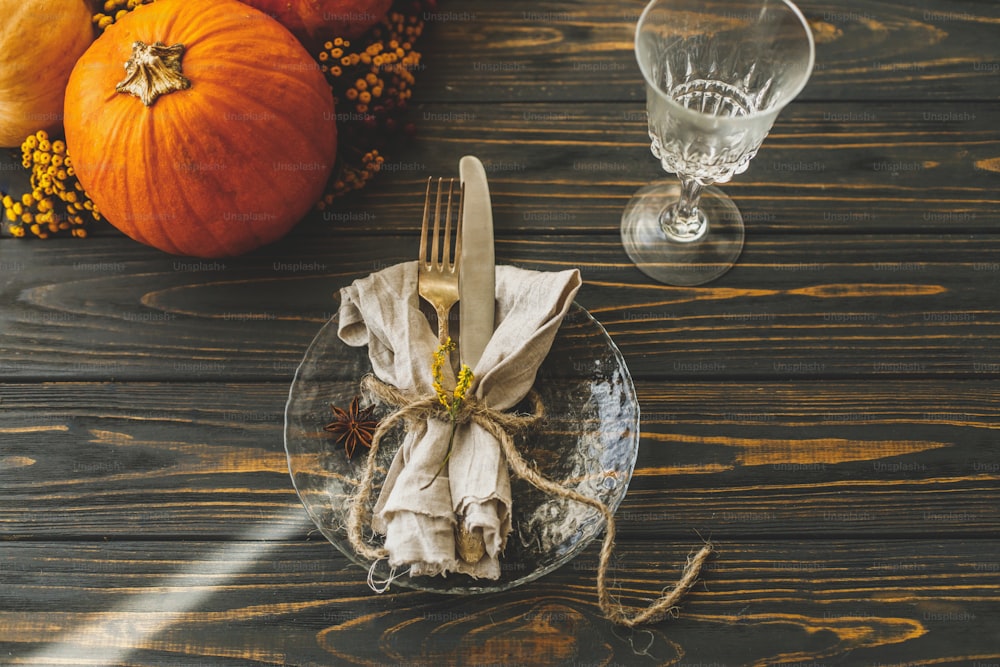 Thanksgiving dinner table setting. Stylish plate with cutlery, linen napkin and pumpkin with autumnal flowers on rustic table. Autumn wedding catering, eco friendly arrangement