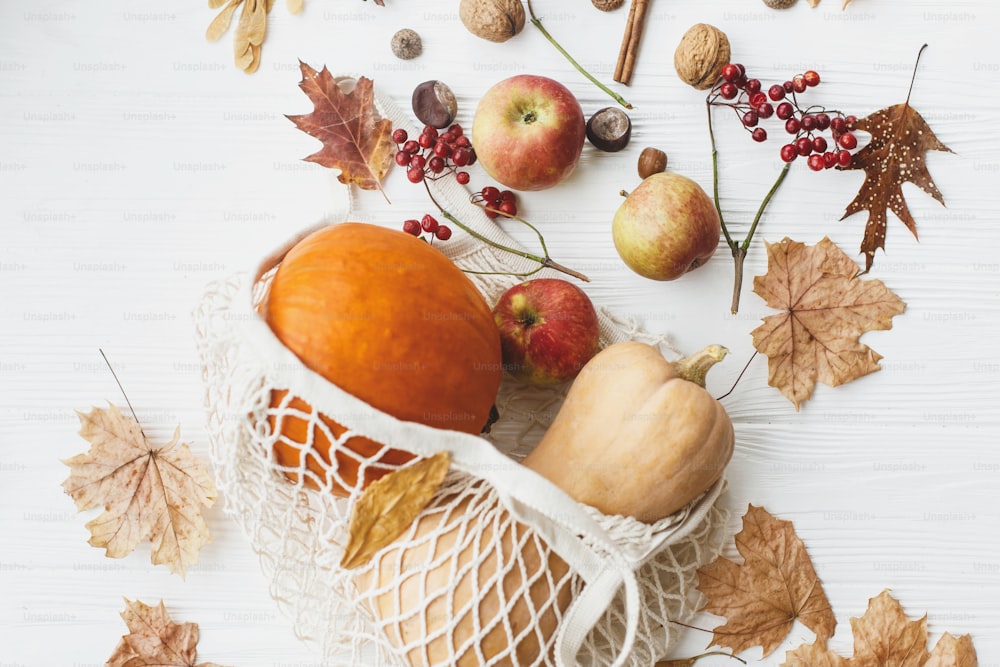 Wiederverwendbare Einkaufstasche mit Kürbissen, Äpfeln, Beeren, Nüssen und Herbstlaub auf weißem Holzgrund. Umweltfreundliches Thanksgiving. Zero Waste Feiertage, Herbsternte