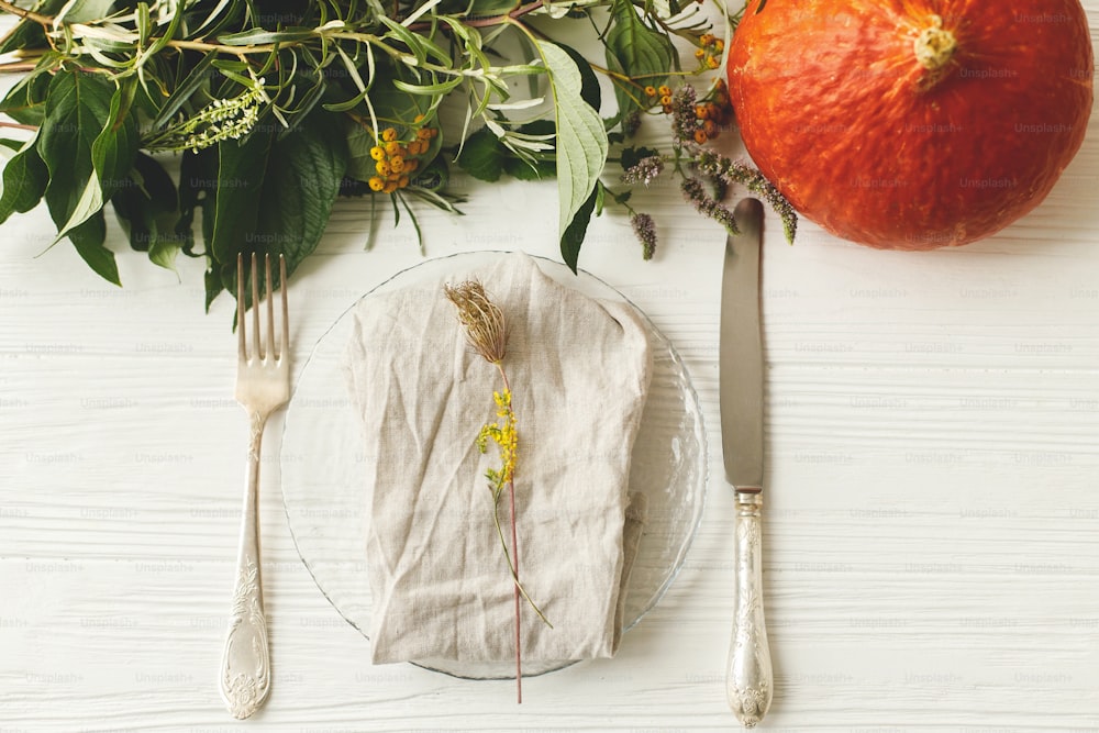 추수감사절 식탁 세팅, 친환경 배치. 흰색 테이블에 칼과 가을 장식, 호박, 천연 가지, 가을 꽃이 있는 세련된 접시. 가을 결혼식