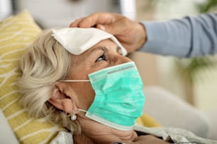 Sick senior woman wearing protective face mask while her husband cools her forehead with a handkerchief.