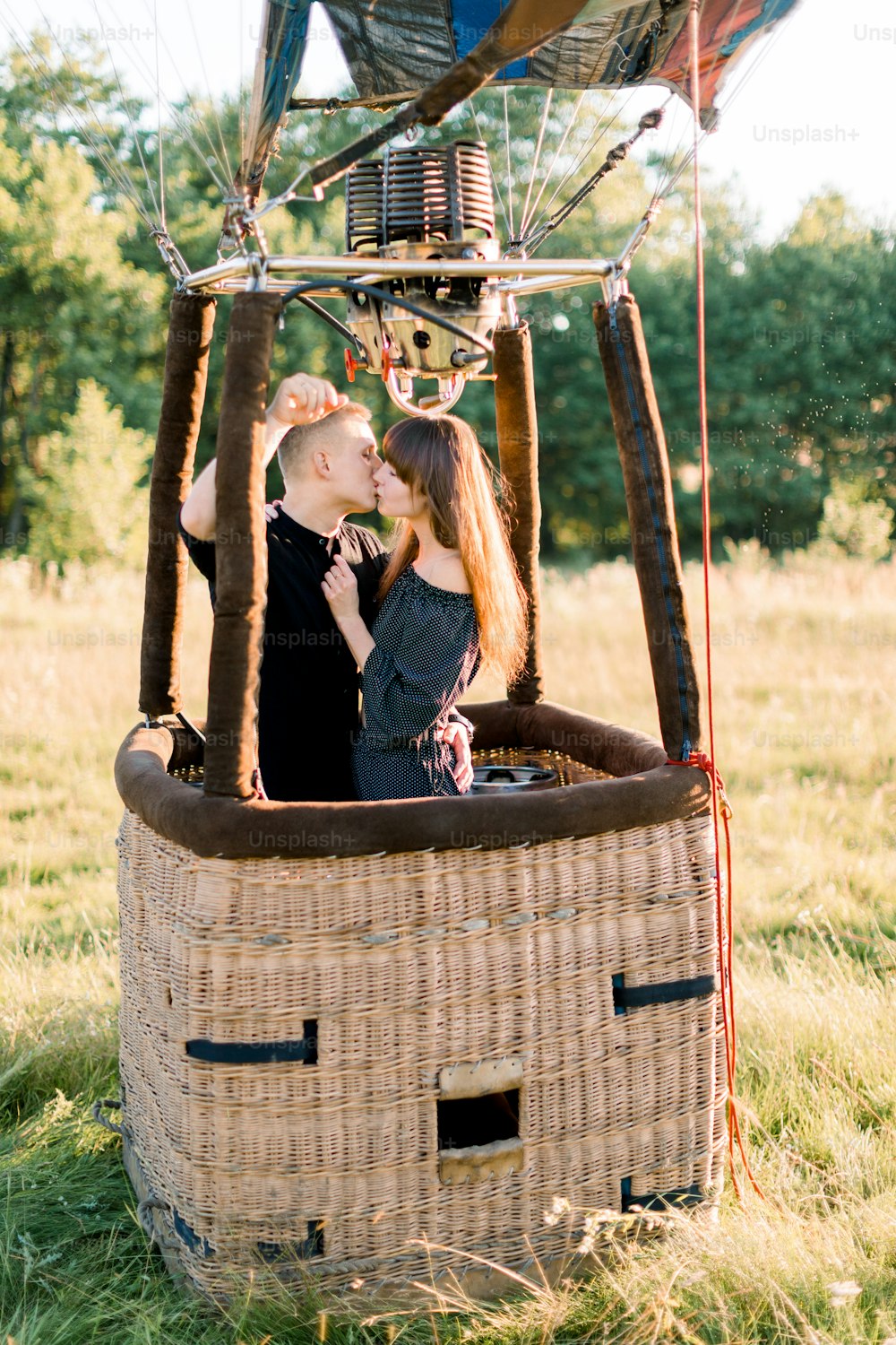 Young beautiful couple in black clothes, kissing in the basket of hot air balloon, enjoying their first fly in warm summer sunrise in the field.
