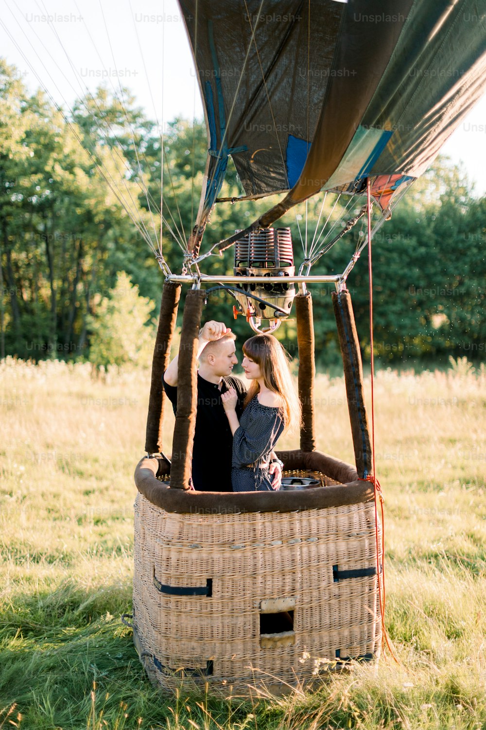 Beautiful young smiling couple in love, wearing black casual clothes, staying and hugging in air balloon basket at sunset, ready for their first balloon flight.