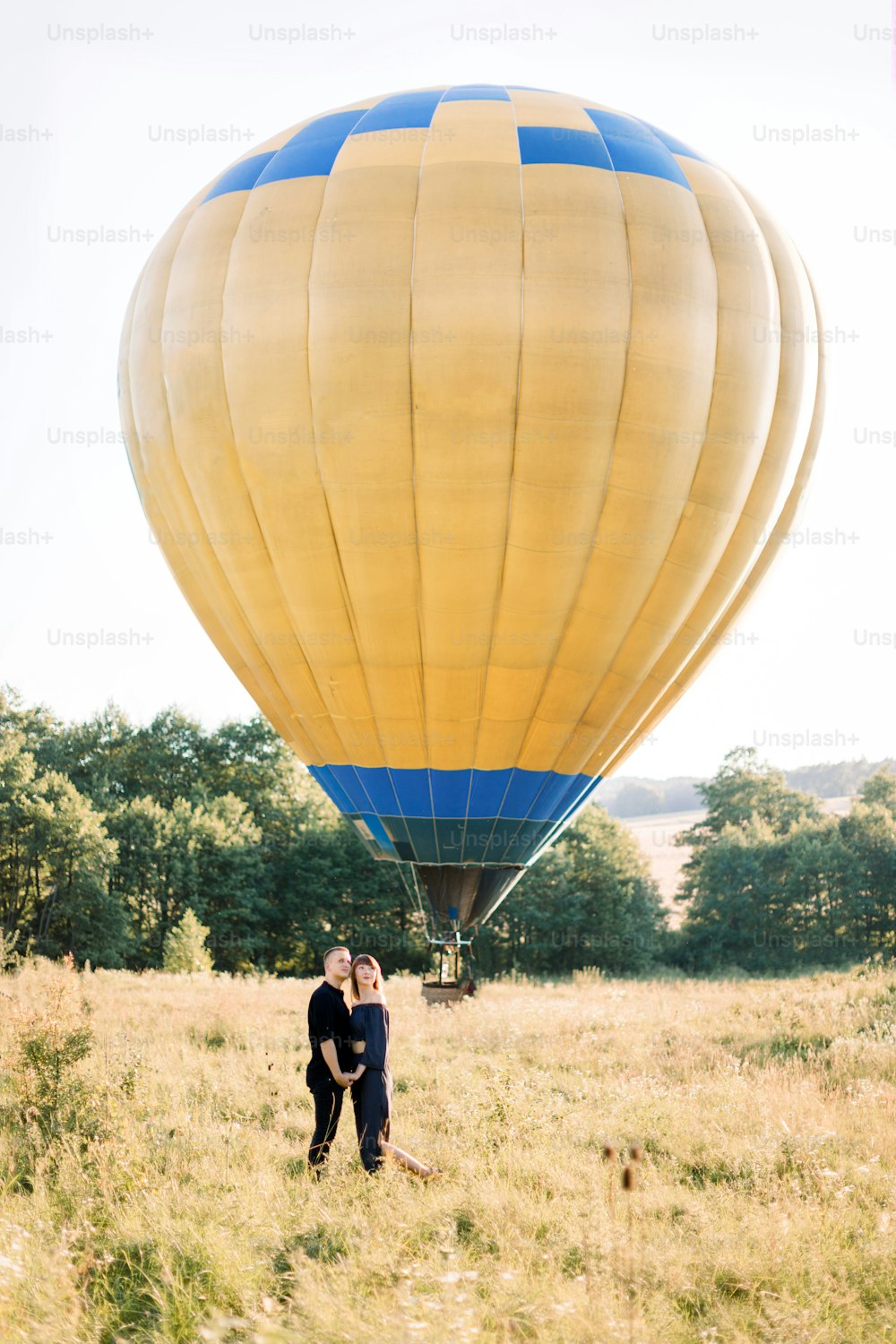 Retrato de cuerpo entero de una encantadora pareja joven vestida de negro, abrazándose y disfrutando de un paseo de verano por el campo, esperando su recorrido en globo aerostático. Globo aerostático amarillo en el fondo.