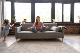 Calm woman concentrating on yoga exercises on couch at home while two noisy kids laughing, running, catching each other, having fun, stress reduction concept.